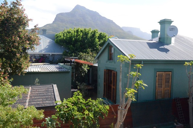 A view of the surrounding buildings, showcasing their green exteriors and distinctive architectural features. Lush trees and plants frame the scene, while a prominent mountain stands majestically in the background under a clear sky.