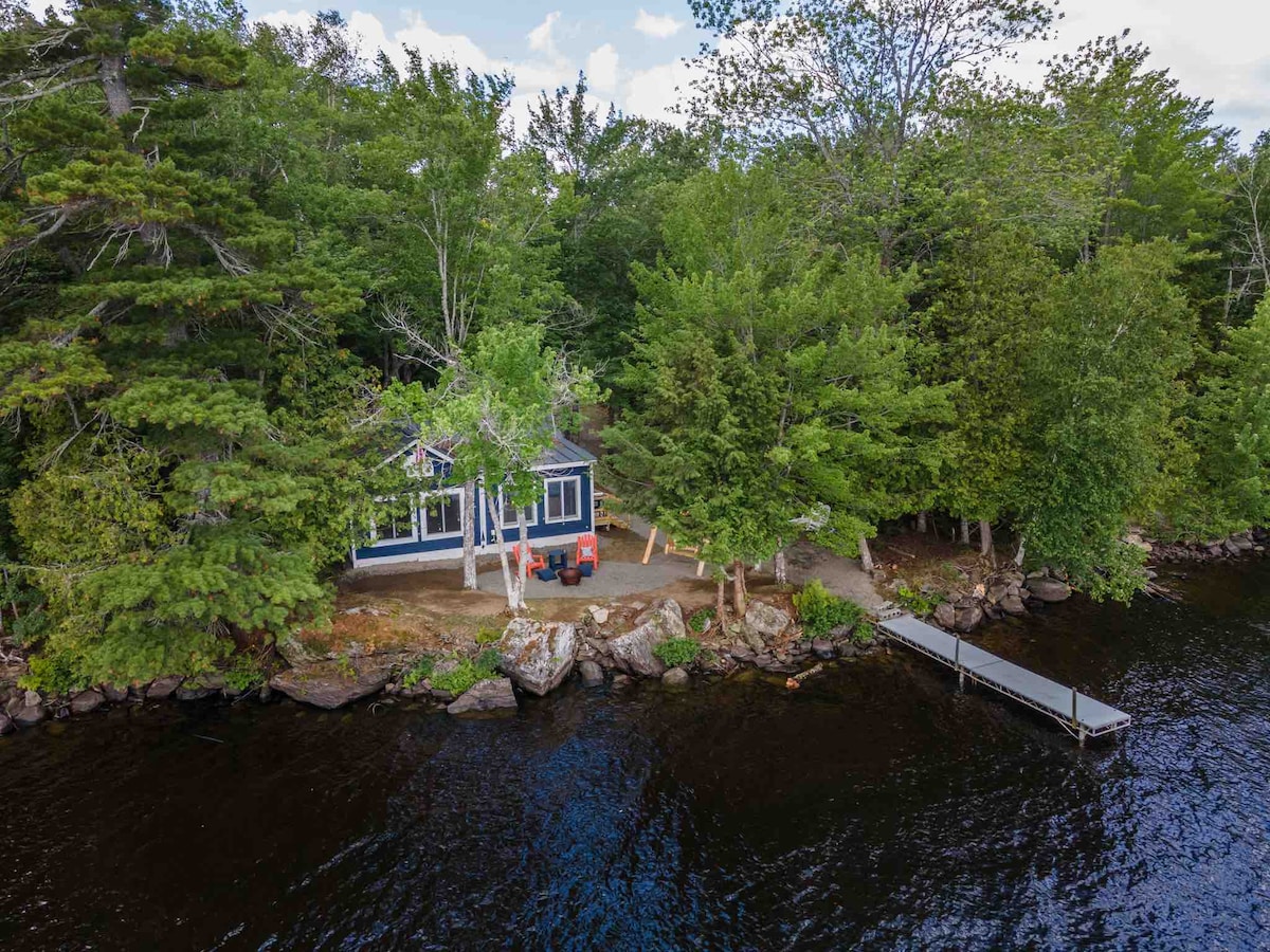 A cabin is situated on the edge of Pushaw Lake, surrounded by lush trees. The outdoor space includes a small dock extending into the water, with a seating area visible on the shore. The cabin's large windows invite natural light, blending the indoor and outdoor environment.