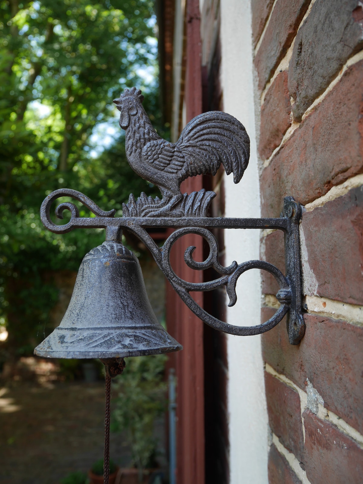 An ornamental cast iron bell is mounted onto a brick wall. The bell features a rooster design perched above it, with intricate detailing in the metalwork. Lush greenery is visible in the background, adding a natural setting to this charming exterior detail.