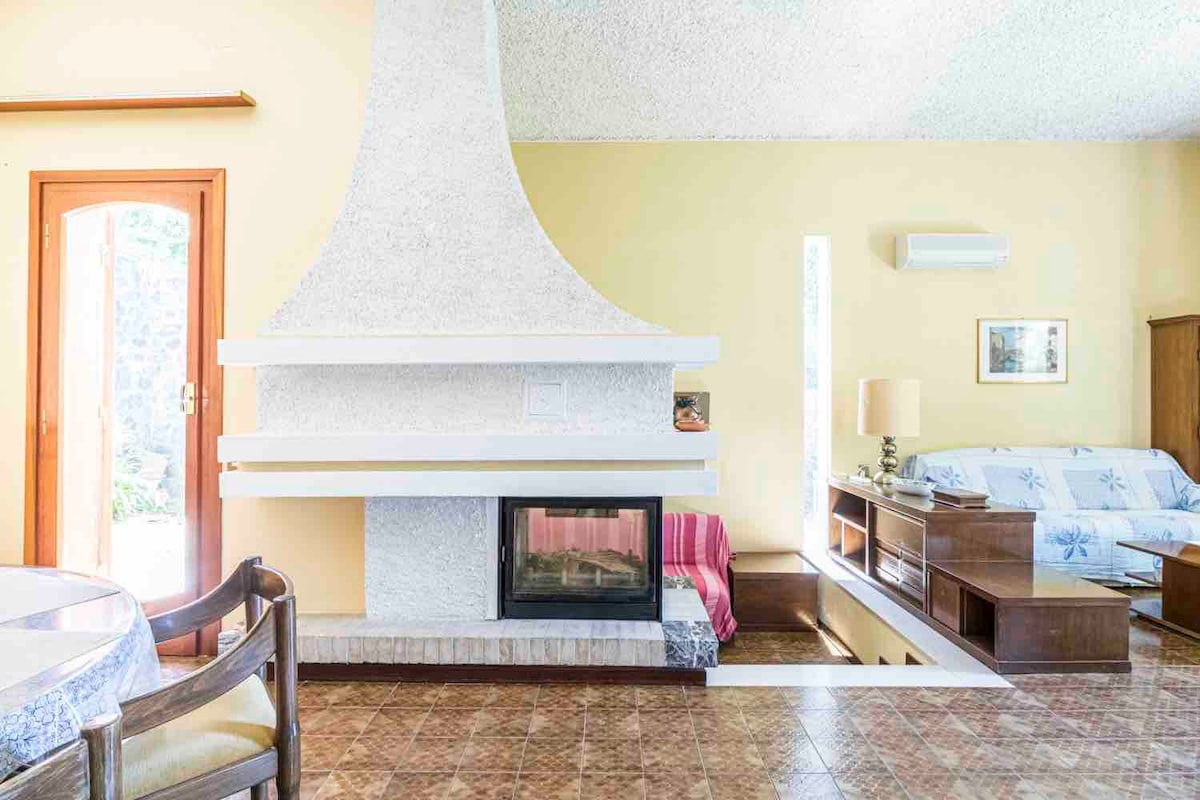 A spacious living area features a unique white fireplace as a focal point. Natural light enters through large windows, illuminating the pastel yellow walls. A comfortable sofa with patterned cushions is positioned beside wooden furniture, while a circular dining table is visible in the foreground.