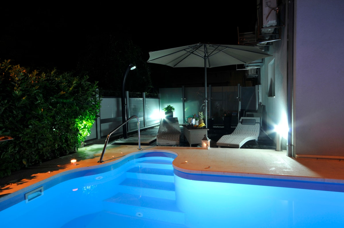 A well-lit outdoor space features a crystal-clear pool with steps leading in, surrounded by lush greenery. A large umbrella shades a seating area with comfortable furniture, illuminated by soft lights, creating a serene evening atmosphere.