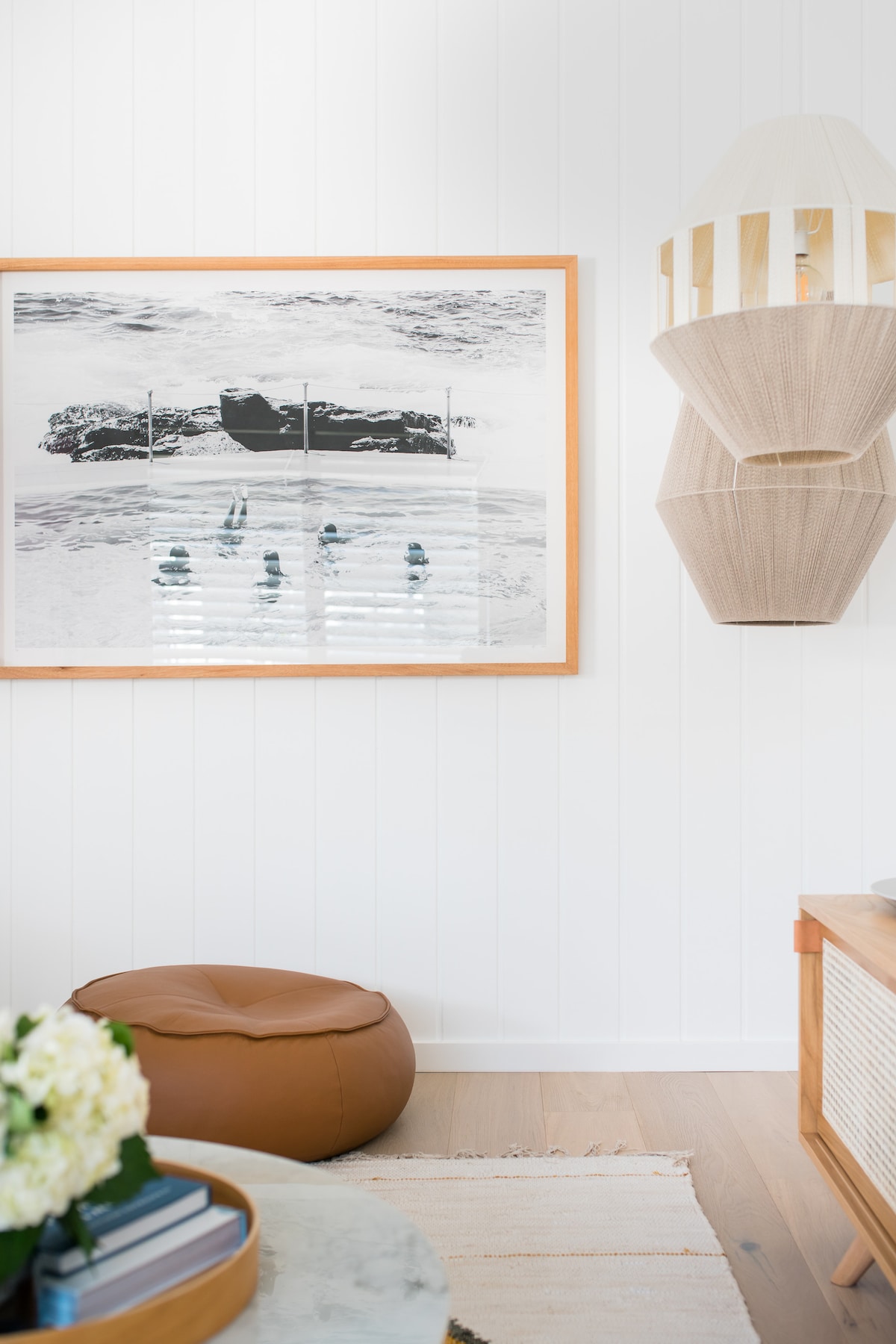 A living area features a monochromatic artwork depicting a beach scene, framed in wood and hung against a white paneled wall. A modern pendant light and a round brown pouf are present, along with a low wooden table topped with decorative items, creating a relaxed atmosphere.