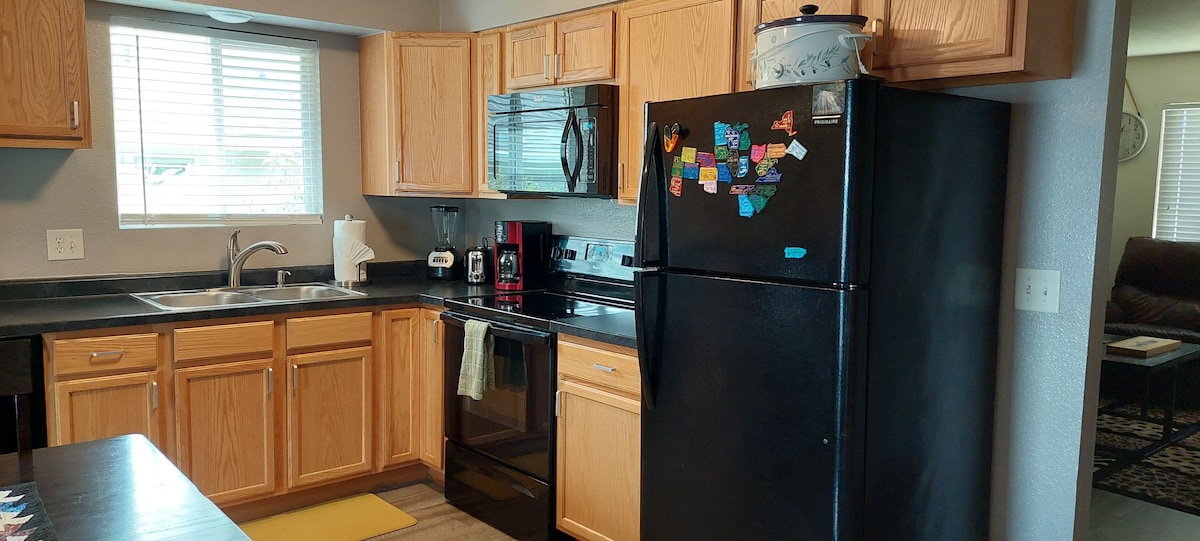 The kitchen features wooden cabinetry, a black refrigerator with notes and magnets on the door, and a black stove and microwave. A window provides natural light, and a yellow mat is placed in front of the sink, enhancing the functional space.