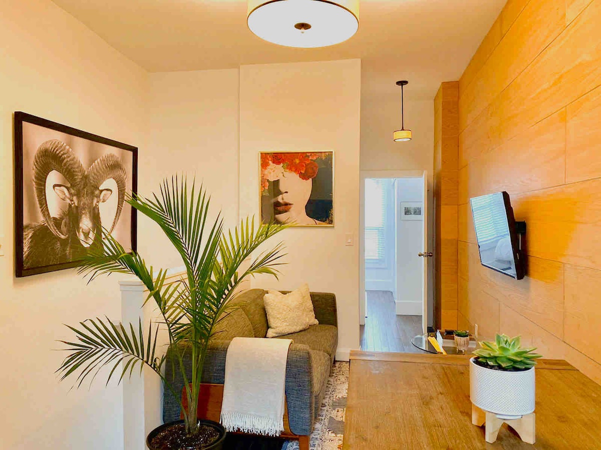 A comfortable living area features a modern couch draped with a soft throw. Artwork decorates the walls, including a striking piece of a woman's face and a framed image of rams. A potted plant adds a touch of greenery, complementing the warm wood accents.