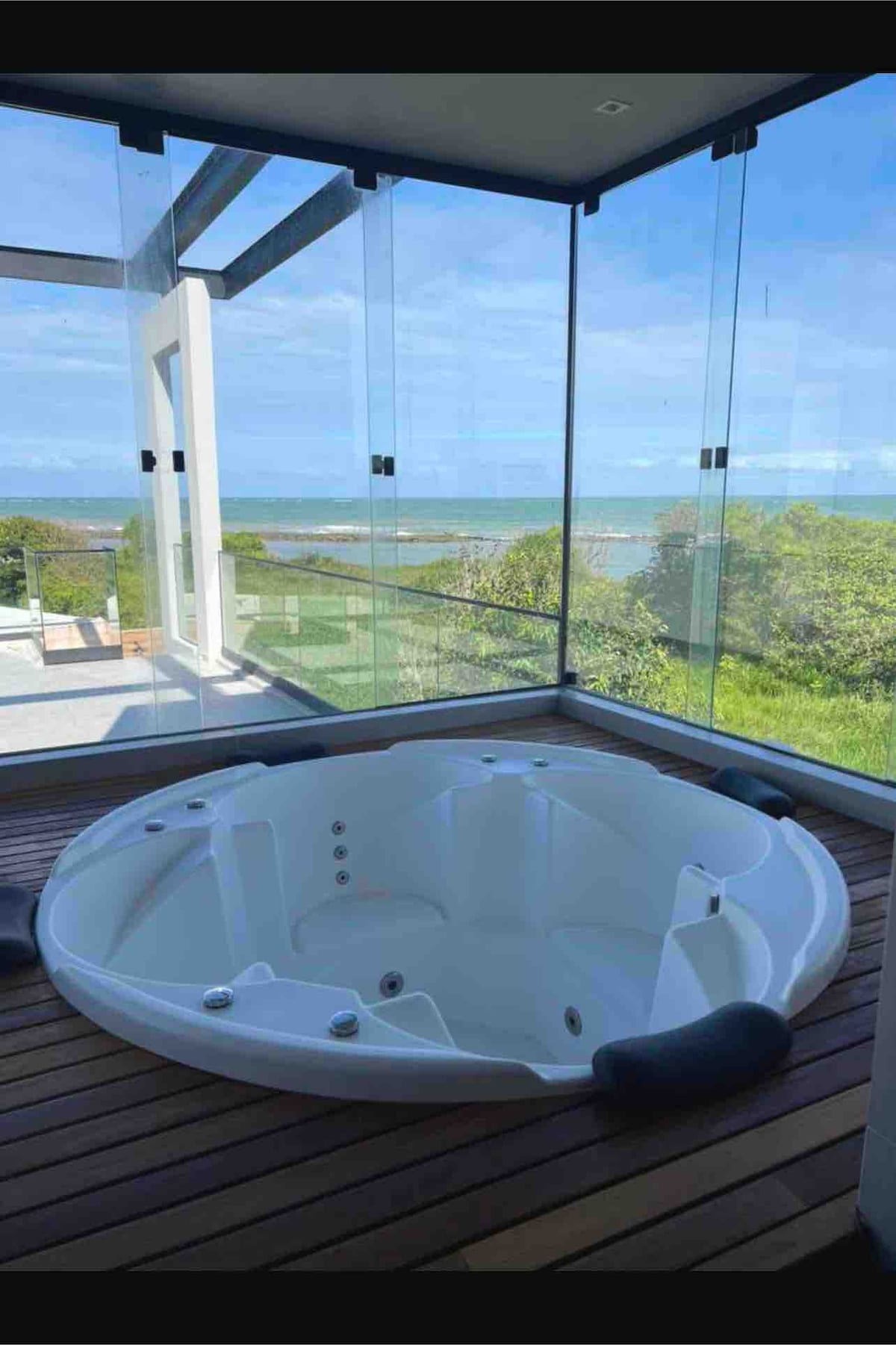 A spacious hot tub is positioned on a wooden deck, surrounded by clear glass walls that provide panoramic views of the sea and greenery. Natural light floods the area, enhancing the serene setting.
