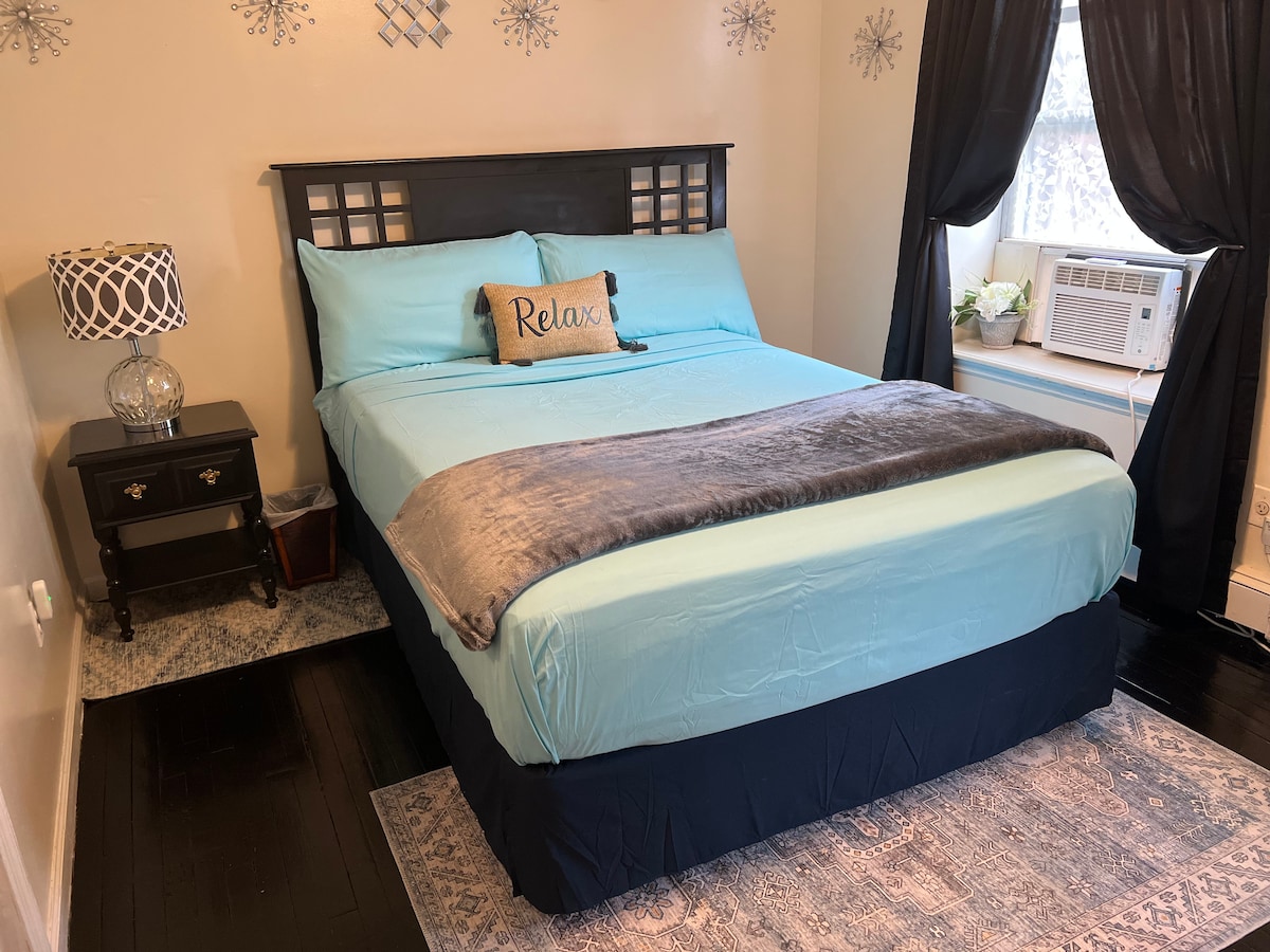 A medium-sized bedroom features a spacious bed dressed in light blue linens and a cozy gray throw. A bedside table with a lamp and decorative pillow is present, alongside a window with dark curtains. An air conditioning unit is visible near the window.