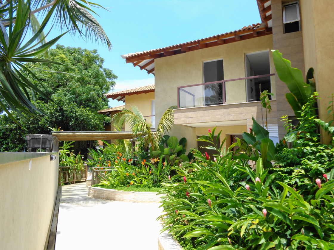 The exterior of the house is surrounded by lush greenery, featuring a variety of tropical plants and flowers. Two stories of the building are visible, with large windows allowing natural light to enter. A pathway leads to the entrance, enhancing accessibility.
