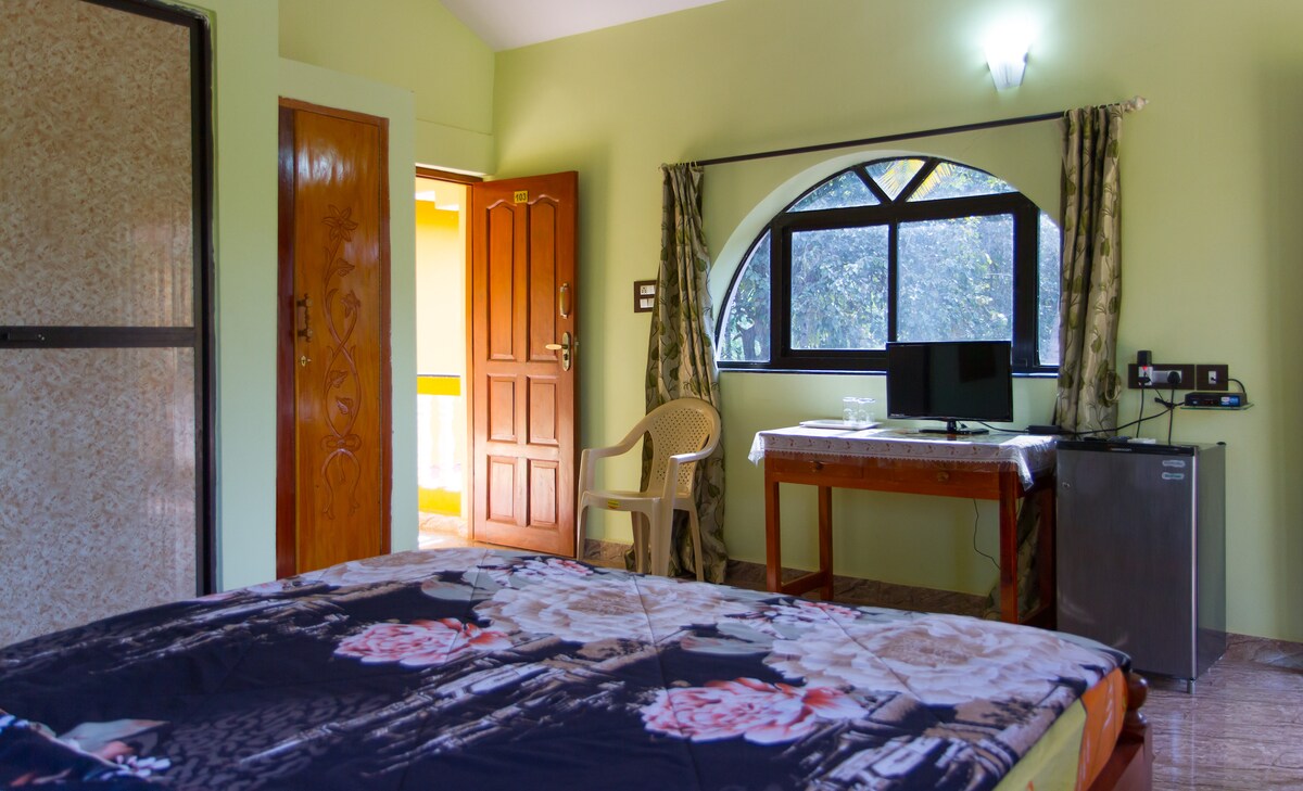 A spacious bedroom features light green walls and a large window allowing natural light. A comfortable bed with floral bedding is positioned next to a writing table and chair. A television is set on the table, and a minibar can be seen to the side.