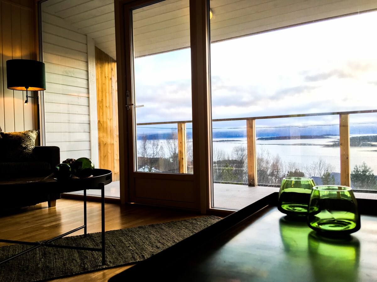 The interior features a cozy seating area with a soft sofa, and a side table displaying green glassware. Large glass doors open to a spacious balcony, offering expansive views of the surrounding fjords and landscapes. Natural light fills the room through the open doors.