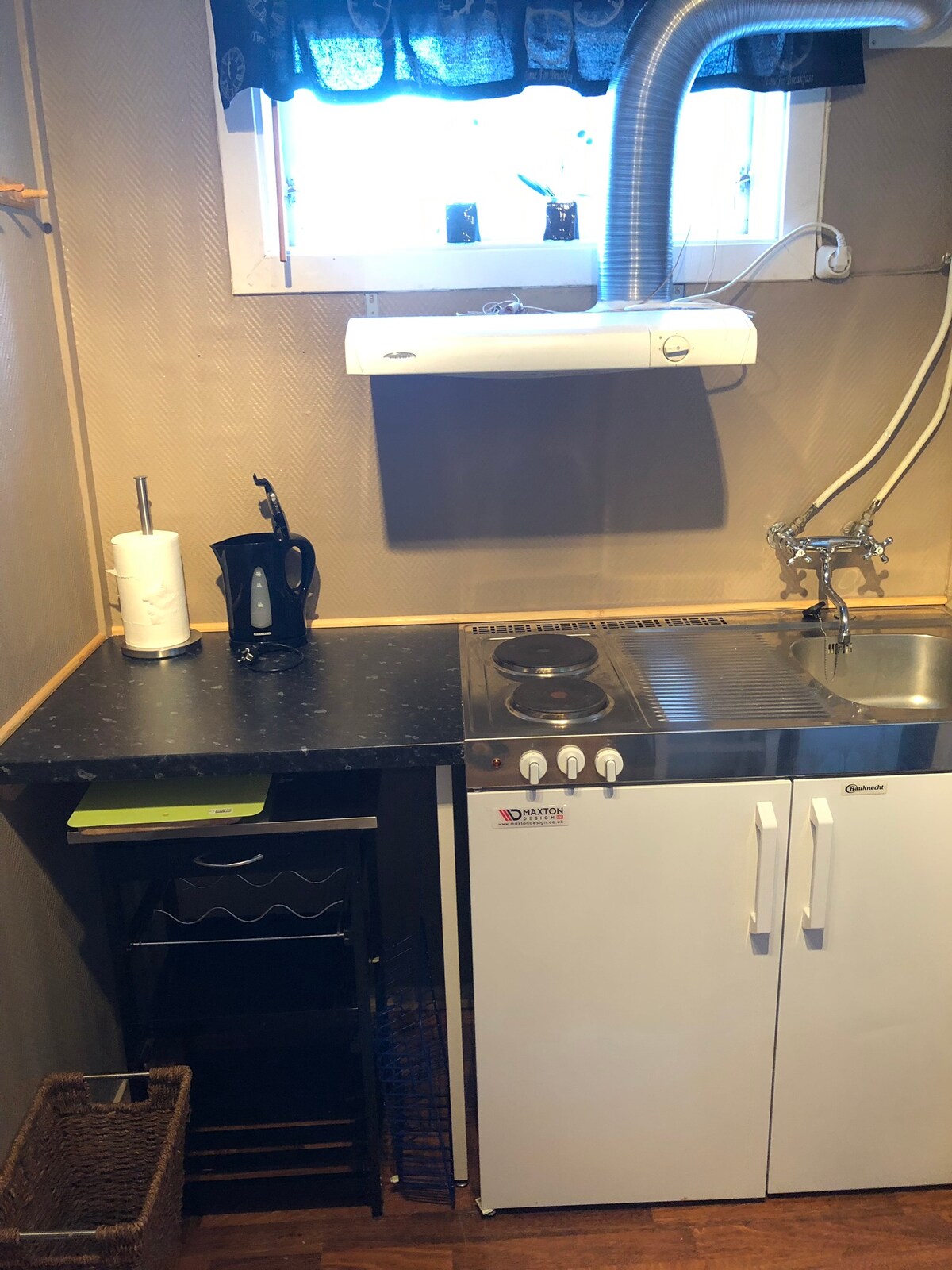 A compact kitchenette is displayed, featuring a black countertop with a coffee maker, a small cooktop, and a sink. Below, a white cabinet offers storage space. Light filters in through a window adorned with a blue curtain, enhancing the functional layout.