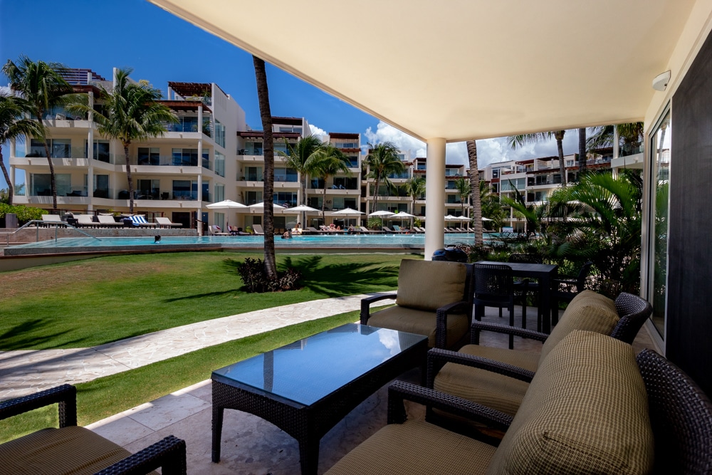 An outdoor terrace features comfortable rattan seating and a glass-top table, overlooking a well-maintained lawn and a spacious infinity pool. Palm trees provide shade, while the modern buildings of the property are visible in the background under a clear blue sky.