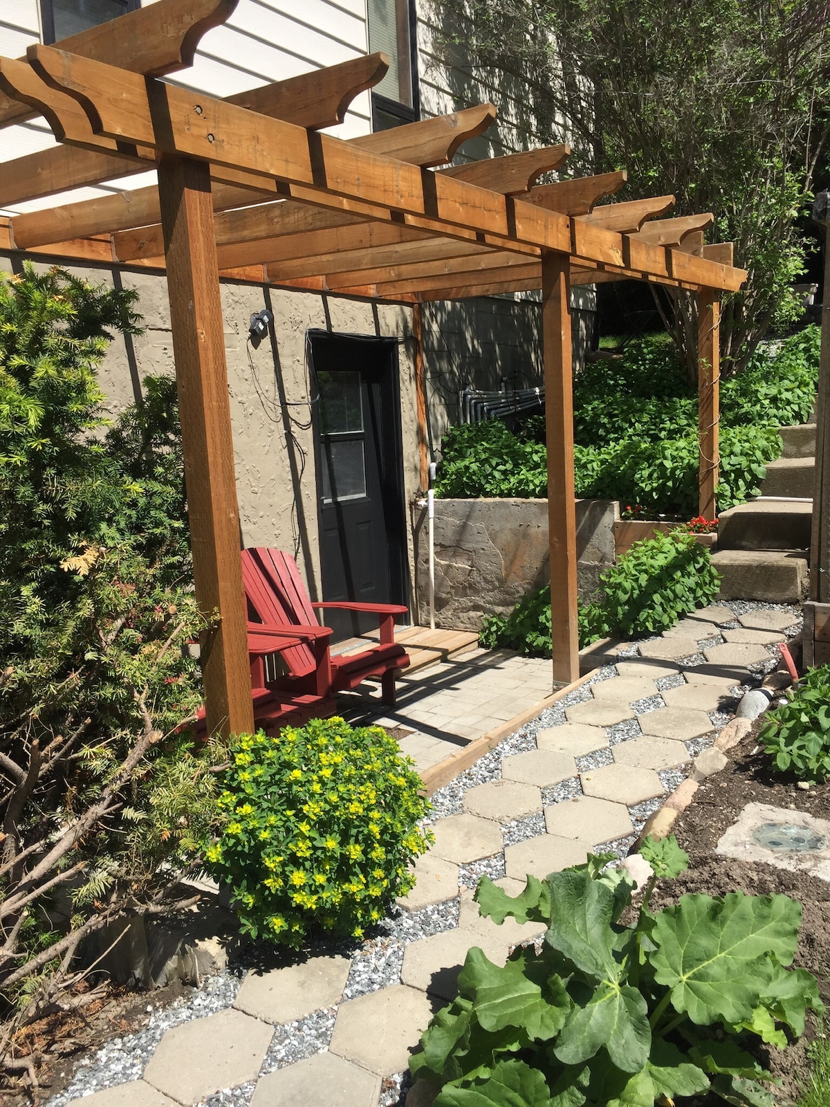 An inviting entryway features a wooden pergola with trellis, complemented by a pair of red adirondack chairs. Lush greenery surrounds the pathway, which is lined with stone pavers and edged with flowering plants, leading to the entrance of the suite.