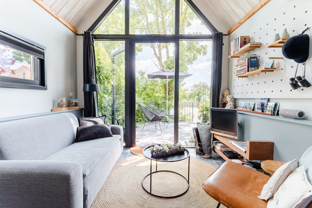 A contemporary living space is characterized by large glass doors that open to a tranquil garden. A comfortable gray sofa sits opposite a small TV and a stylish coffee table. Natural light fills the room, with decorative shelves displaying various books and art.