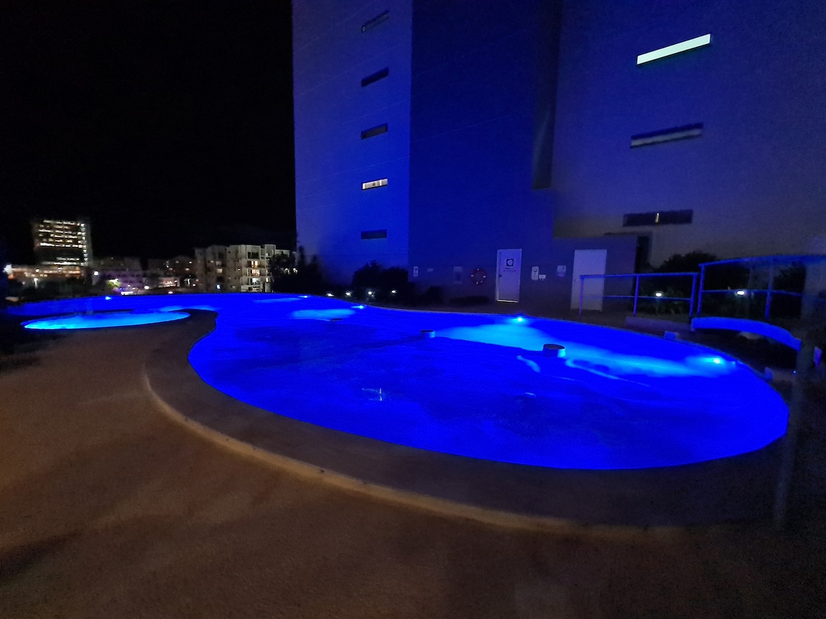 The outdoor pool area is illuminated with soft blue lighting, creating a soothing ambiance at night. The pool features unique shapes and is surrounded by a clean, paved deck, providing a tranquil space for relaxation.