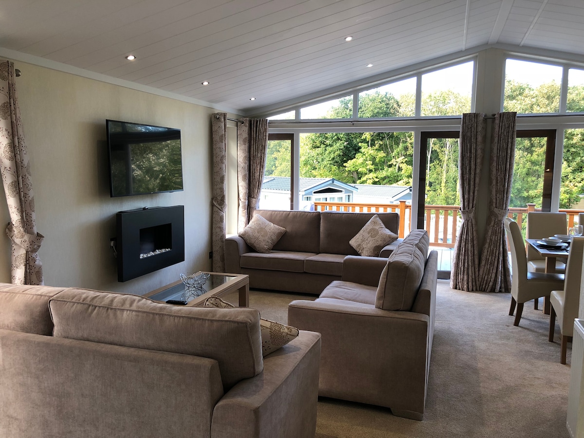 A spacious open-plan living area features large windows, allowing natural light to illuminate the neutral-toned furnishings. Three comfortable settees are positioned around a wall-mounted television, with a stylish fireplace below. The adjoining dining area is visible, enhancing the inviting layout.
