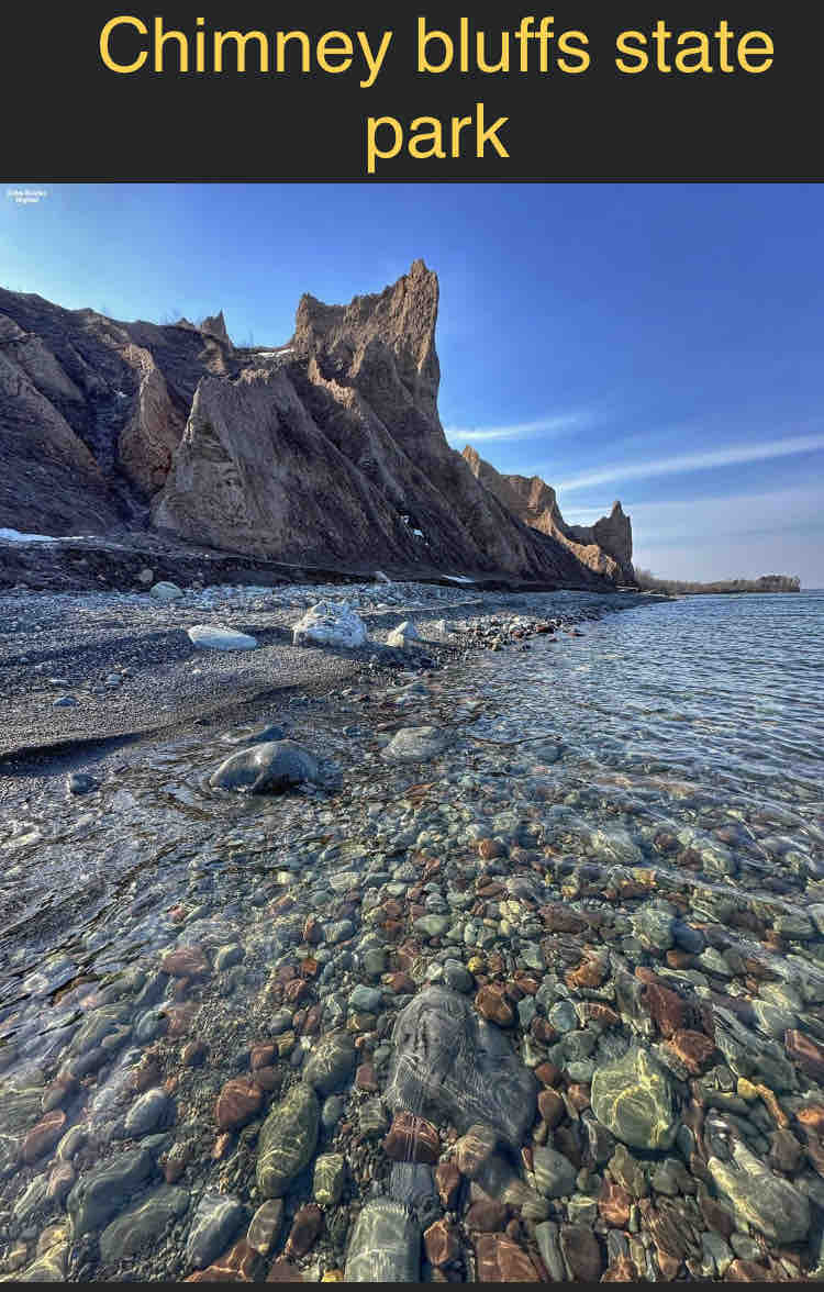 Lush rock formations rise majestically above a tranquil shoreline. Clear waters reveal colorful stones resting in the gentle surf, while the clear blue sky and distant cliffs create a serene backdrop.