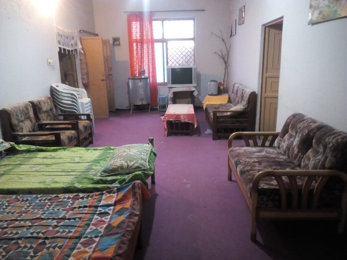 A spacious living area features multiple seating options, including two patterned sofas and several chairs. Colorful throws cover two beds, providing additional lounging space. Natural light enters through a window with sheer curtains, illuminating the carpeted floor.