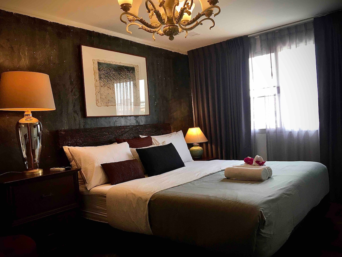 A welcoming bedroom features a spacious bed with crisp white linens and a soft gray throw, complemented by accent pillows. A decorative chandelier hangs from the ceiling above, while natural light enters through a large window, framed by sheer and dark curtains. A bedside lamp offers soft illumination.