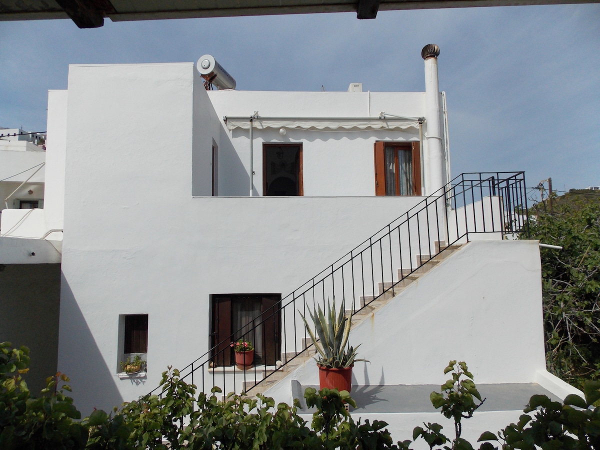 The exterior of a two-story white building is visible, featuring a staircase that leads to an upper level. Windows with wooden frames and small potted plants are seen, along with a simple railing and an outdoor smoking chimney. Green foliage surrounds the base, providing a natural contrast.