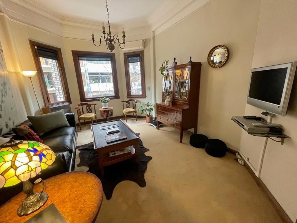 A living room is depicted, featuring a dark leather sofa and two vintage wooden chairs positioned around a wooden coffee table. A decorative cabinet stands against one wall, with a television mounted nearby. Natural light enters through multiple windows, accompanied by a decorative lamp on a side table.