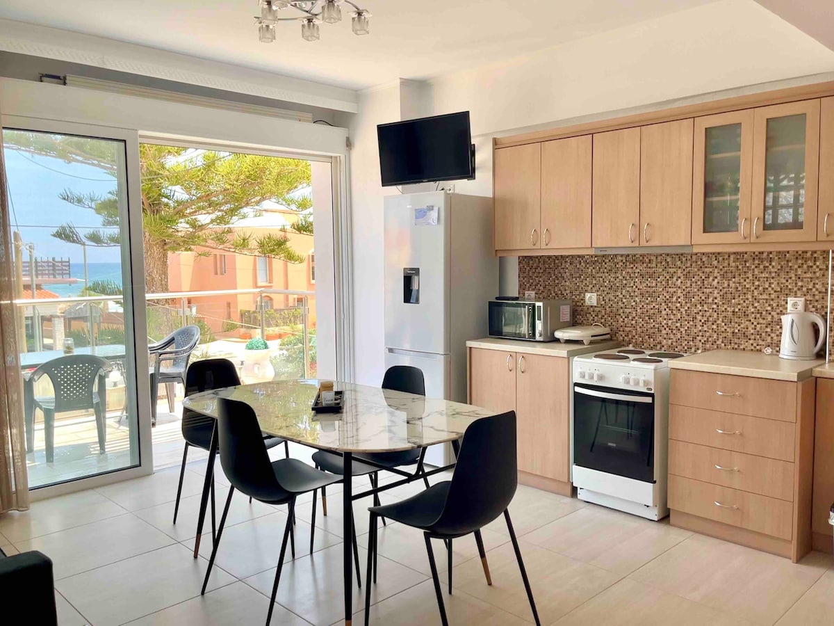 A modern kitchen and dining area features a marble-top table surrounded by six black chairs. Large glass doors allow natural light and views of the outdoor space. Kitchen appliances include a refrigerator, microwave, and stove, with wooden cabinetry and a tiled backsplash providing a clean aesthetic.