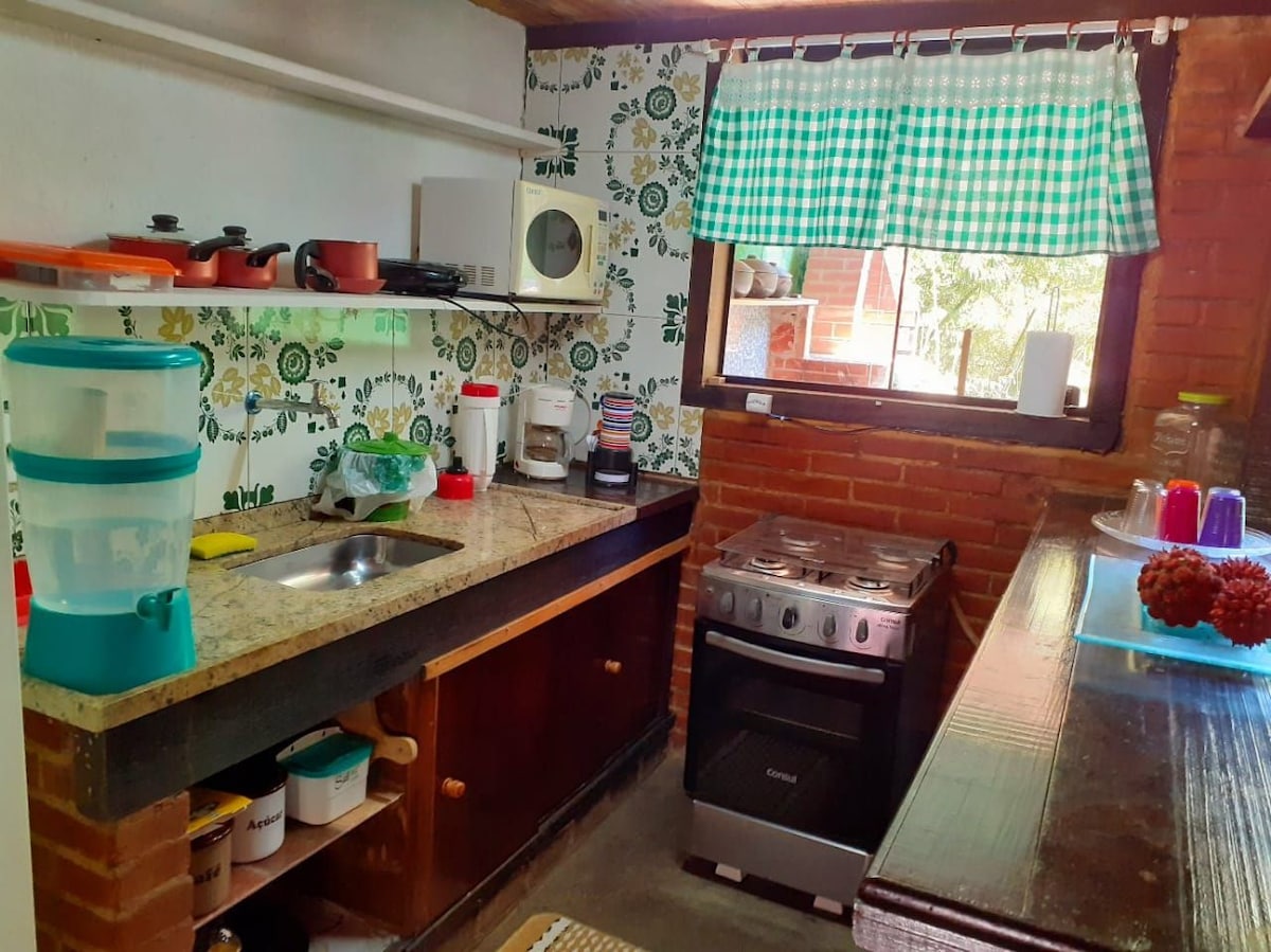 The kitchen features a granite countertop, equipped with a stainless-steel sink and a gas stove with an oven. Shelves hold kitchenware and a small appliance collection. A patterned wallpaper adds character, while natural light filters through a window with green checkered curtains.