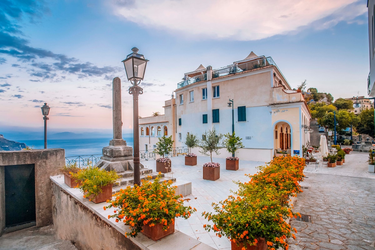 A charming plaza is depicted, featuring decorative planters filled with vibrant orange flowers. A stone column stands prominently alongside street lamps. In the background, white buildings with blue shutters overlook the serene sea, with gentle colors of the sunset visible in the sky.