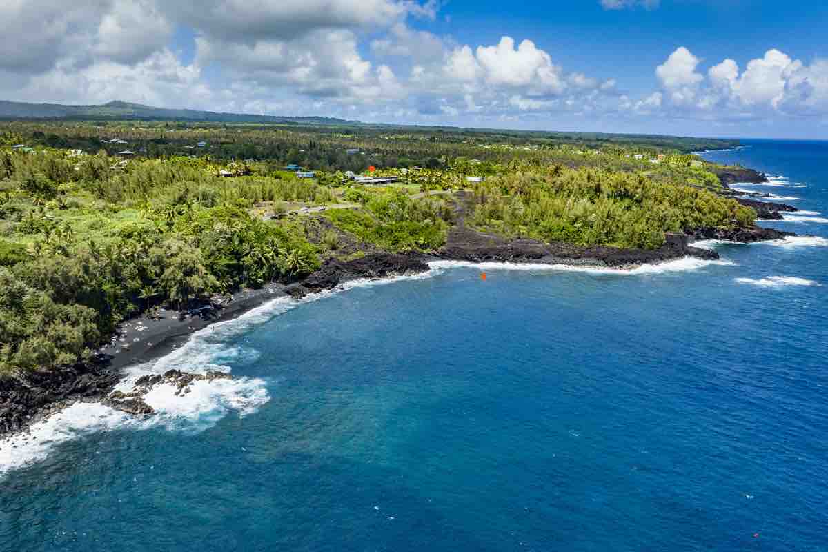 An aerial view captures the rugged coastline where gentle waves meet the serene black sand beach. Lush greenery and volcanic rock outline the shore, while vibrant blue waters reflect the sky. The coastline is framed by distant hills dotted with trees, creating a peaceful natural landscape.