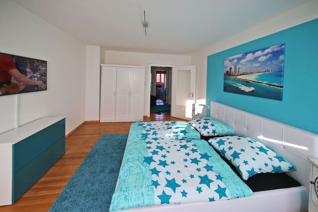 Schlafzimmer mit bequemen Doppelbett in wunderschönem Miami Türkis. Mit TV und Sideboard sowie einer Sitzbank