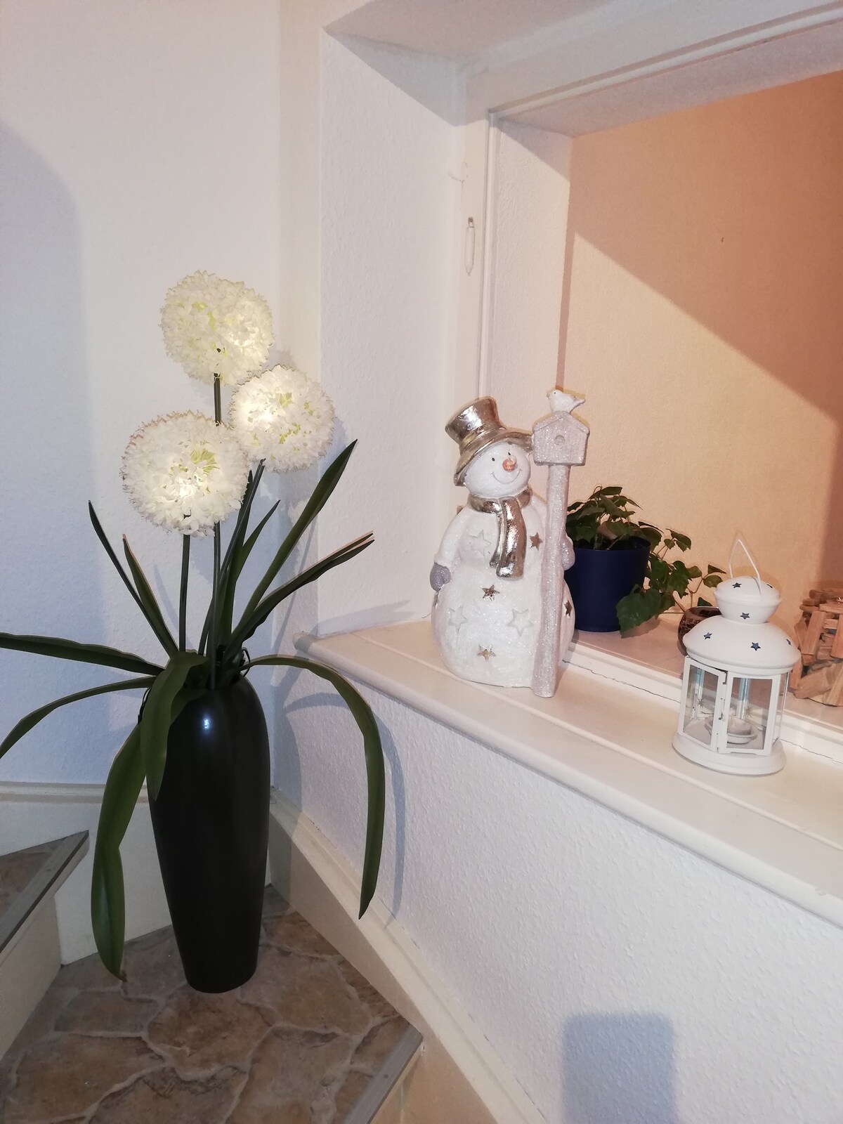 A decorative corner features a tall black vase with white flowers alongside a cheerful snowman figurine wearing a hat and holding a star. A small lantern and a potted plant are also visible on the windowsill, enhancing the cozy ambiance.