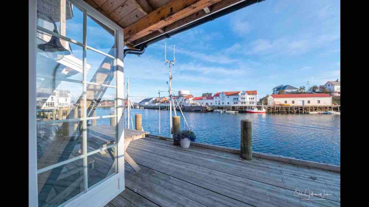A spacious deck extends over the water, offering views of boats docked in the harbor. Light wooden planks form the floor, while blue skies create a bright atmosphere. Potted plants add a touch of greenery, enhancing the waterfront setting.