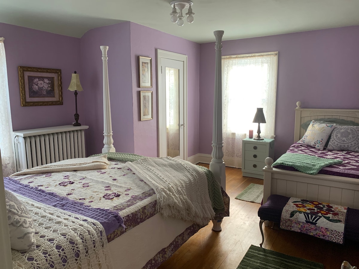 A well-lit bedroom features two beds, one with a decorative quilt and one adorned with a colorful knitted throw. A charming nightstand with a lamp and a window with sheer curtains enhance the inviting atmosphere. Soft pastel walls and wooden flooring create a cozy setting.