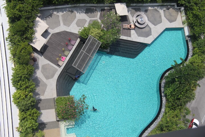 The aerial view captures a serene swimming pool surrounded by landscaped greenery. Sun loungers and shaded seating areas are visible along the pool's edge, offering spaces for relaxation. The pool's turquoise water reflects the sun, inviting a refreshing escape.