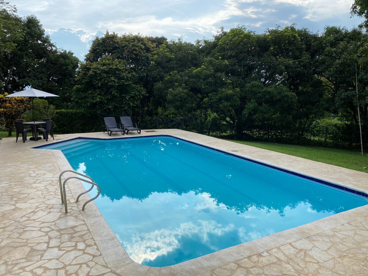 A clear blue swimming pool is framed by a stone patio and surrounded by lush greenery. Two lounge chairs are positioned next to the pool, with a table and umbrella in the shaded area nearby. The tranquil setting invites relaxation under the sky.