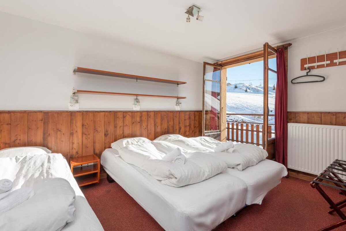 The room features three spacious beds adorned with white linens. A wooden wall provides a warm contrast to the light-colored furnishings. Natural light enters through a large window, with a scenic view of snow-covered slopes outside. A shelf hangs above the beds for storage.
