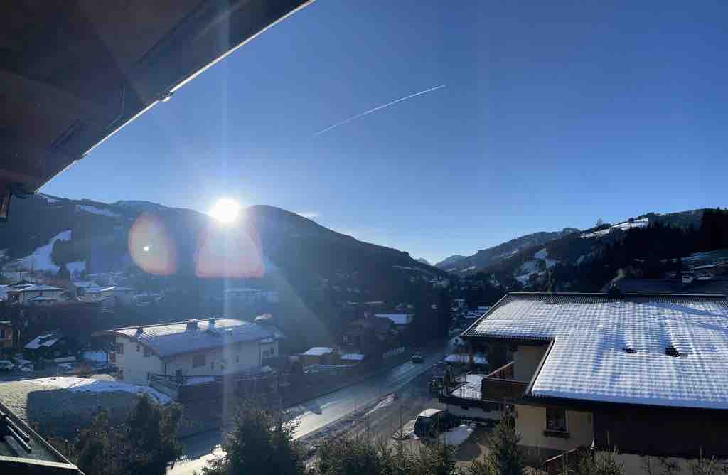 A panoramic view showcases snow-covered mountains under a clear blue sky. The rising sun casts soft light across the landscape, illuminating houses with snow-dusted rooftops in the foreground. The serene atmosphere is enhanced by the untouched white landscape.