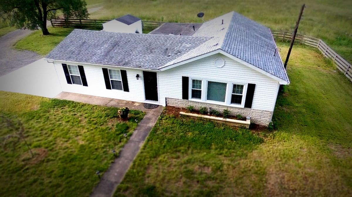 An aerial view of the newly remodeled farmhouse reveals a white exterior with black shutters. A stone-accented wall is visible beneath the windows, while a walkway leads to the entrance. A spacious lawn surrounds the home, bordered by a fence and greenery.