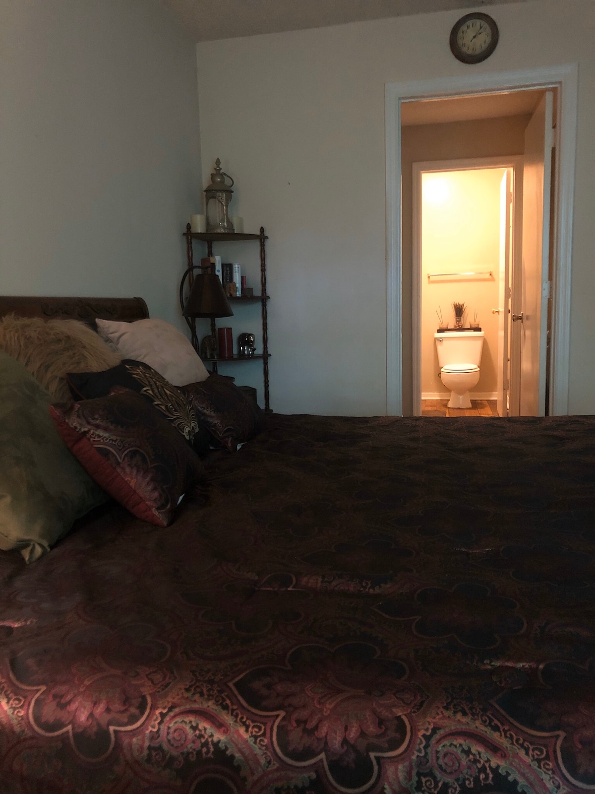 A spacious bed is adorned with decorative pillows and a rich patterned bedspread. A small bookshelf is positioned nearby, showcasing various decorative items. A doorway leads to a well-lit bathroom area, featuring a toilet and additional amenities.