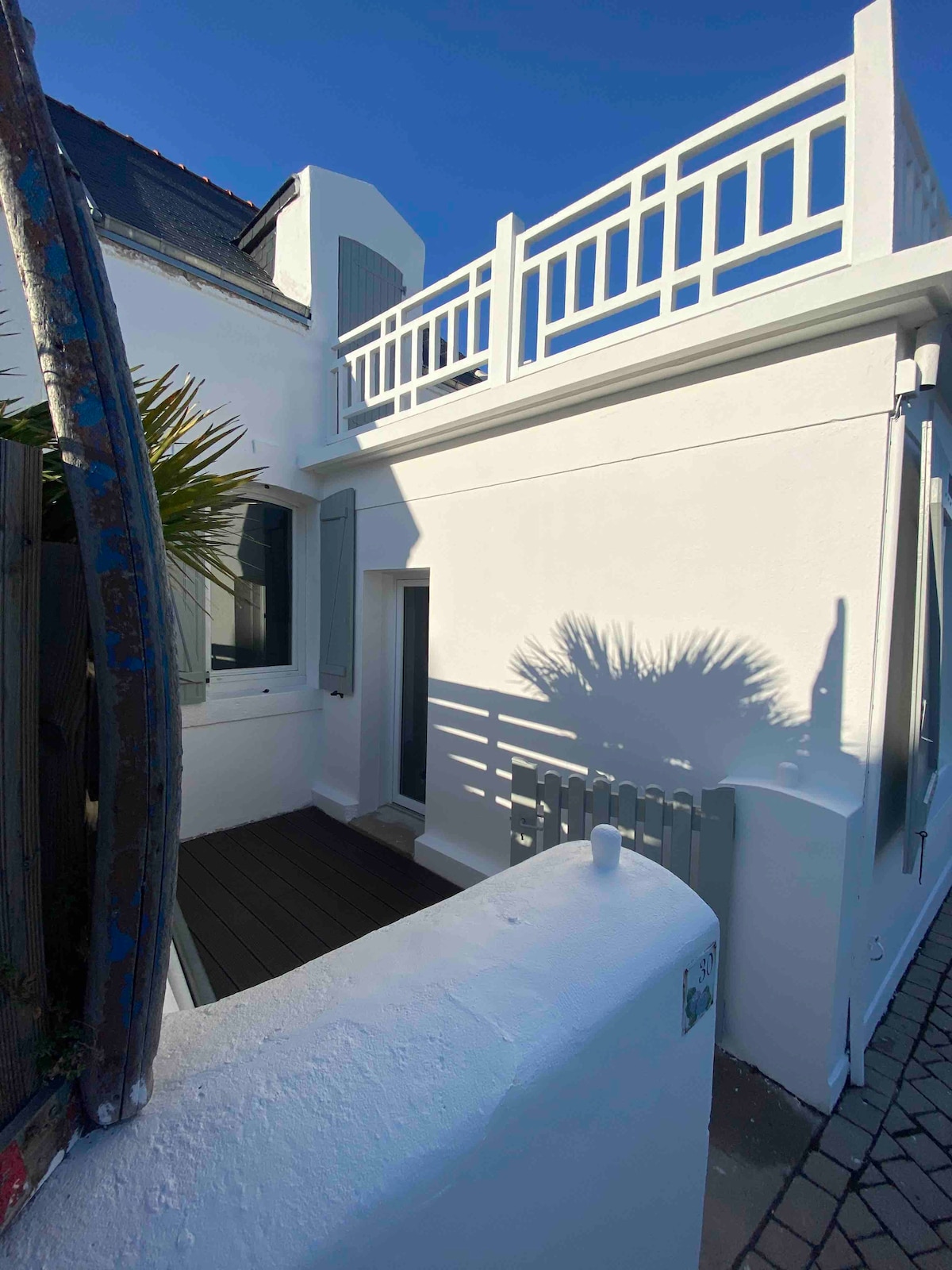 A white exterior of the house is illuminated by sunlight, showcasing a terrace with railing. A shadow of a palm leaf can be seen on the wall, adding texture. The entrance to the house is located at the side, complemented by wooden decking.