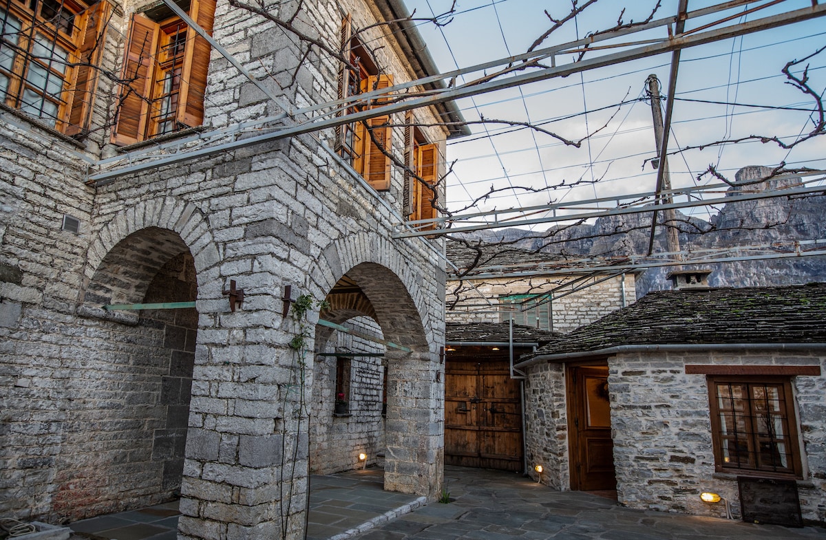 The exterior features a traditional stone structure characterized by arched entrances and wooden shutters. A trellis overhead supports vines, adding to the rustic charm. Natural stone flooring extends throughout the courtyard, with nearby walls showcasing the building's architectural details.