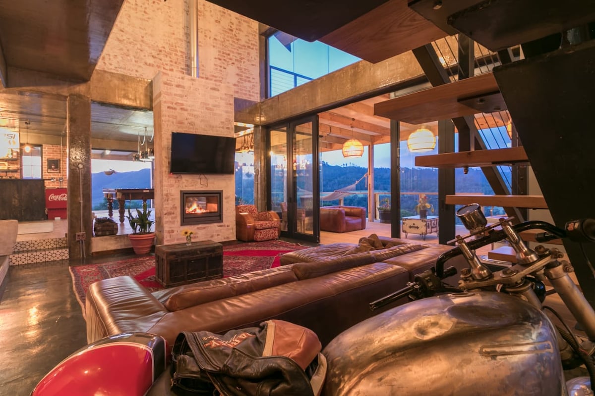An inviting living area features a spacious leather sofa, with a rustic coffee table positioned in front of a modern fireplace. Large glass doors open to reveal panoramic views of the surrounding landscape, with warm light from stylish hanging lamps illuminating the space.