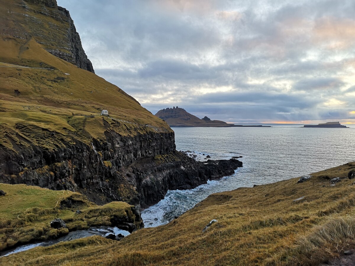 A rugged coastal landscape is depicted, showcasing steep cliffs covered with golden grass. Calm waters reflect the overcast sky, while a distant island formation enhances the scenic view. A small structure is positioned along the coastline, blending harmoniously with the natural surroundings.