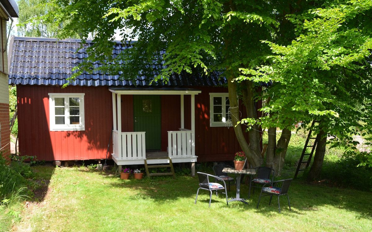 A charming red cottage is nestled among lush greenery, featuring a green door and white-framed windows. A small patio area with a table and chairs is set on the lawn, providing a peaceful spot to relax amidst nature.