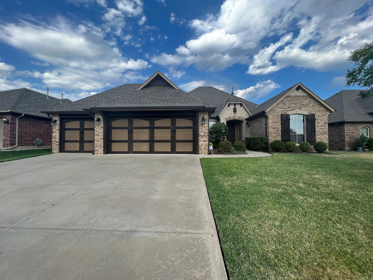A single-story brick home is presented, featuring a spacious driveway and a three-car garage with distinct wooden doors. The neatly trimmed lawn and low shrubs enhance the inviting exterior. Light clouds drift across a blue sky, creating a serene atmosphere.