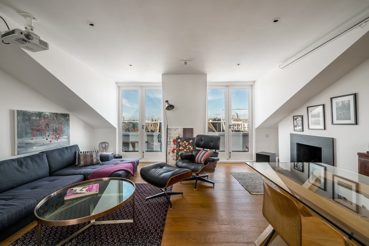 A bright living area features a large, L-shaped sofa facing a glass coffee table. A sleek black lounge chair sits beside a floor lamp, and a glass dining table is positioned nearby. Panoramic windows offer views of the outdoor terrace and cityscape.