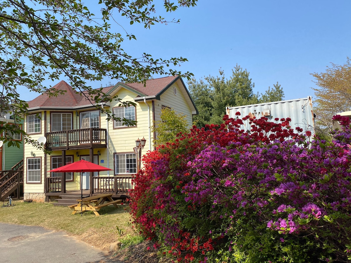 A two-story house with a yellow exterior features a covered balcony and two large windows on the upper level. Vibrant azaleas in shades of pink and purple surround a wooden picnic table underneath a red umbrella, adding a touch of color to the outdoor setting.