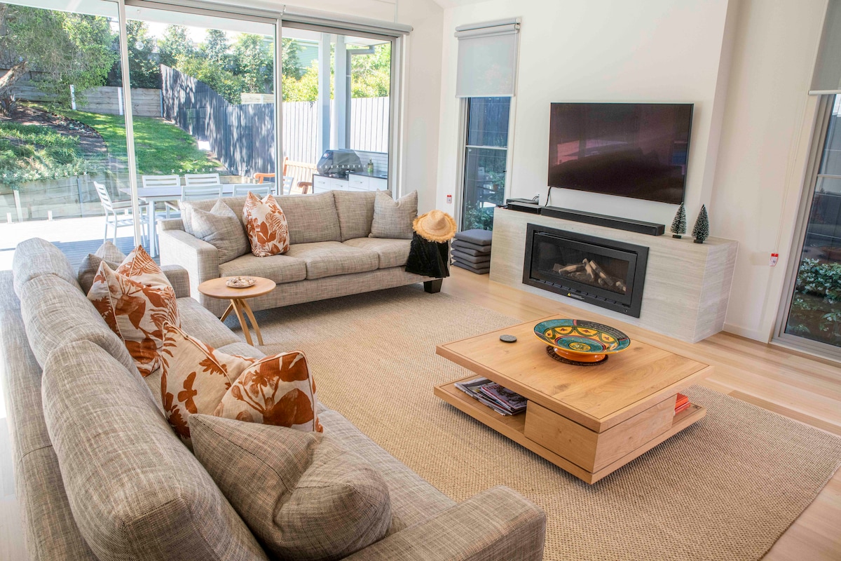 An open-plan living area features two light gray sofas adorned with patterned cushions. A coffee table made of natural wood holds decorative items. A flat-screen TV is mounted on the wall, while large glass doors reveal an outdoor space with seating and a BBQ.