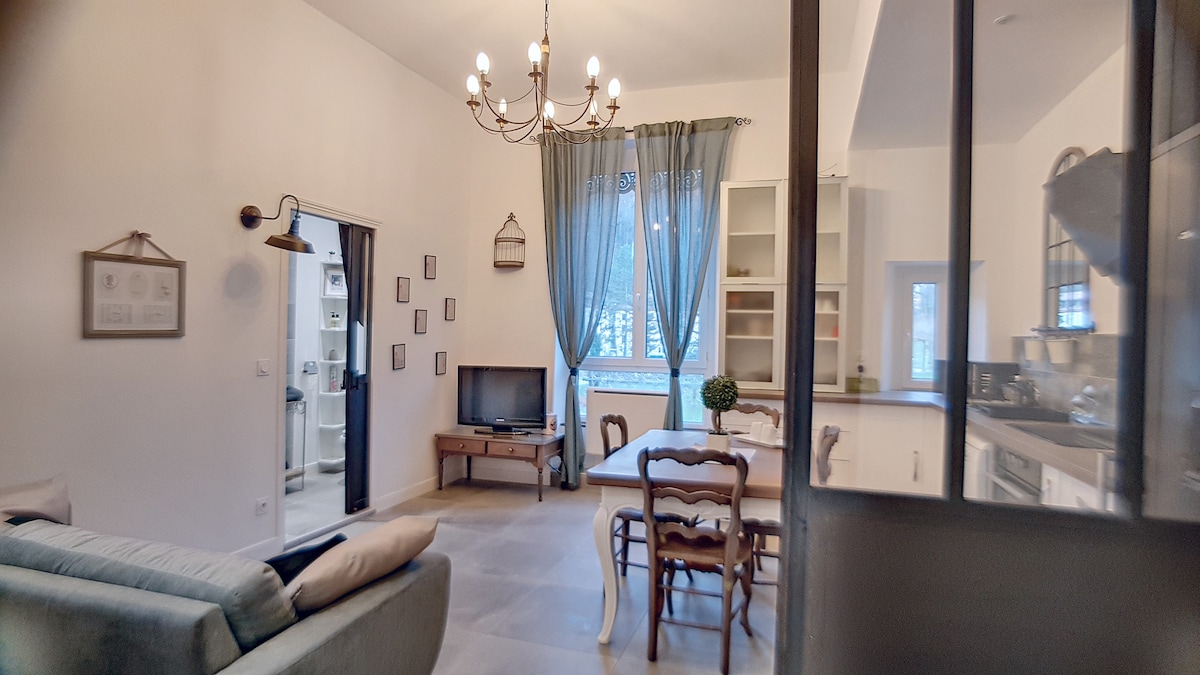 A modern living space is presented with soft-colored walls and a mix of furniture styles. A chandelier hangs from the ceiling. Natural light filters through large windows dressed with light curtains, illuminating a seating area with a sofa and a wooden dining table accompanied by four chairs.