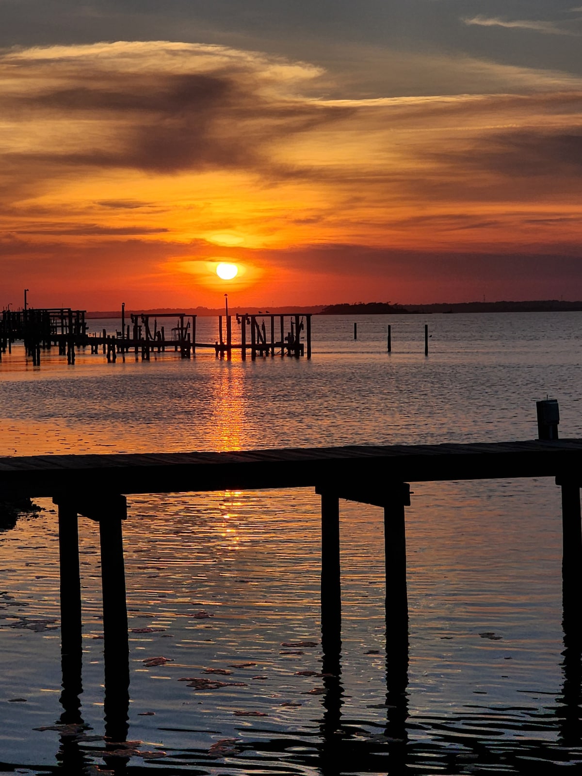 A vibrant sunset casts warm hues across the water, reflected in its gentle ripples. Silhouetted dock pilings create a serene foreground, enhancing the tranquil atmosphere of Bogue Sound, while soft clouds add depth to the colorful sky.