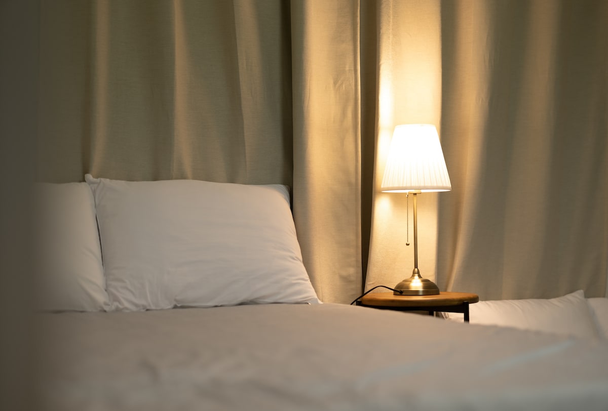 A spacious bed is positioned alongside a nightstand with a softly glowing lamp. The light fabric of the bedding contrasts with the warm tones of the lamp's shade, while muted curtains frame the background, creating a calming atmosphere.